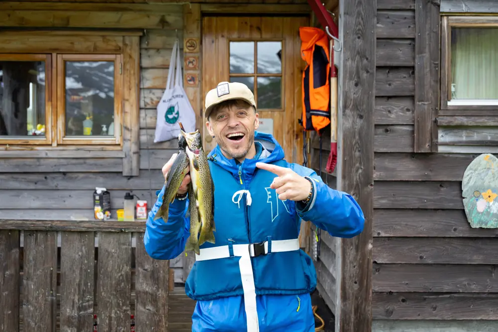Mann i blå jakke smiler foran hytte og peker på noen fjellørret han har fanget.