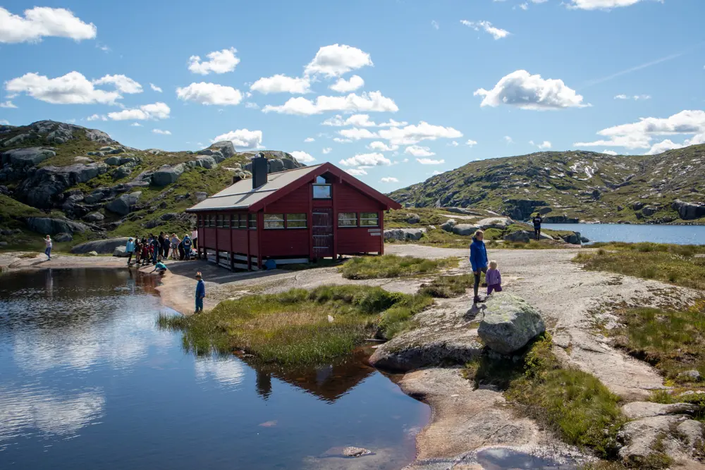 Rød hytte med vann. Barn og voksne utfor hytta. 