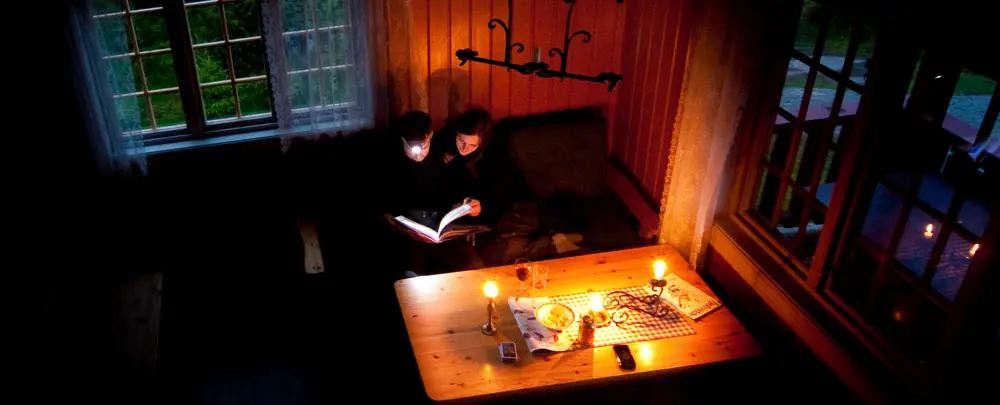 To personer leser en bok sammen i dempet belysning, omgitt av levende lys i en koselig hytte.