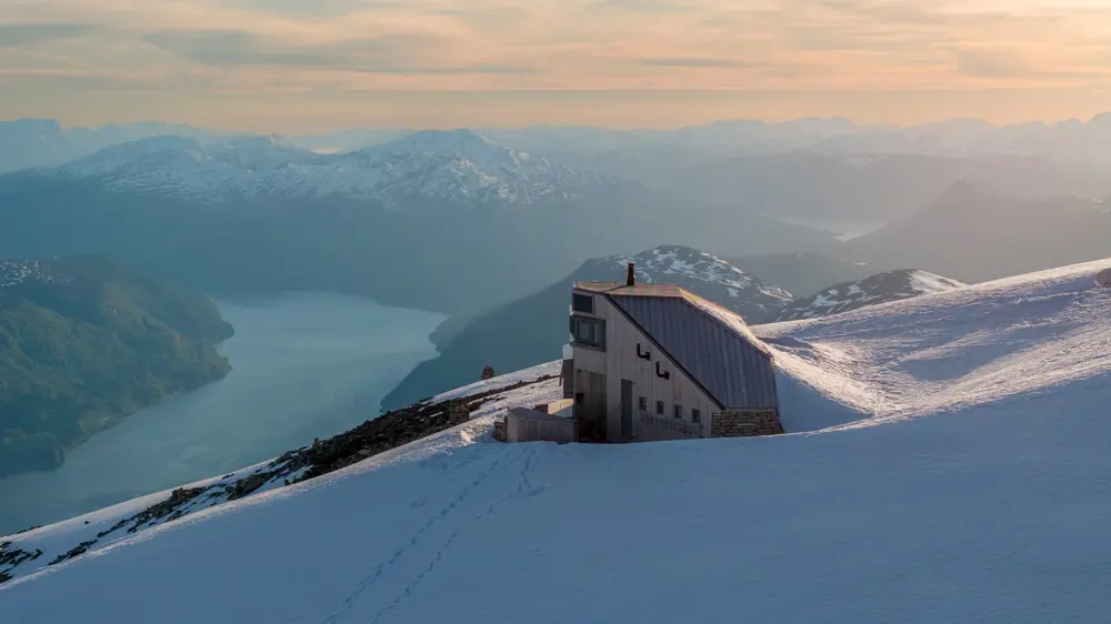 Langstrakt hytte på toppen av fjell i solnedgang med fjord lenger bak.
