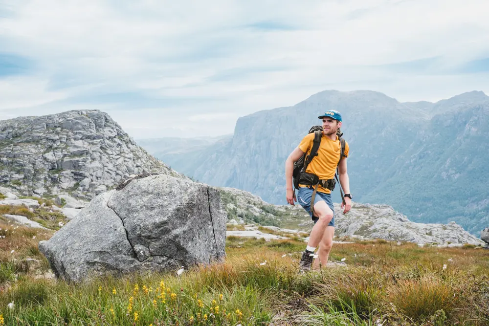 En mann i gul t-skjorte og shorts går på toppen av et fjell.