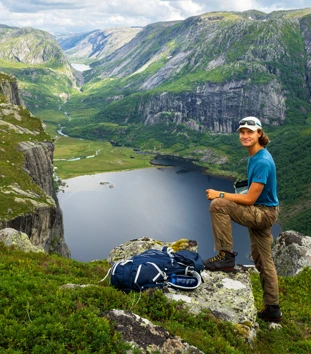 En mann som står på toppen av et fjell med grønt landskap rundt og et vann nederst i bildet.