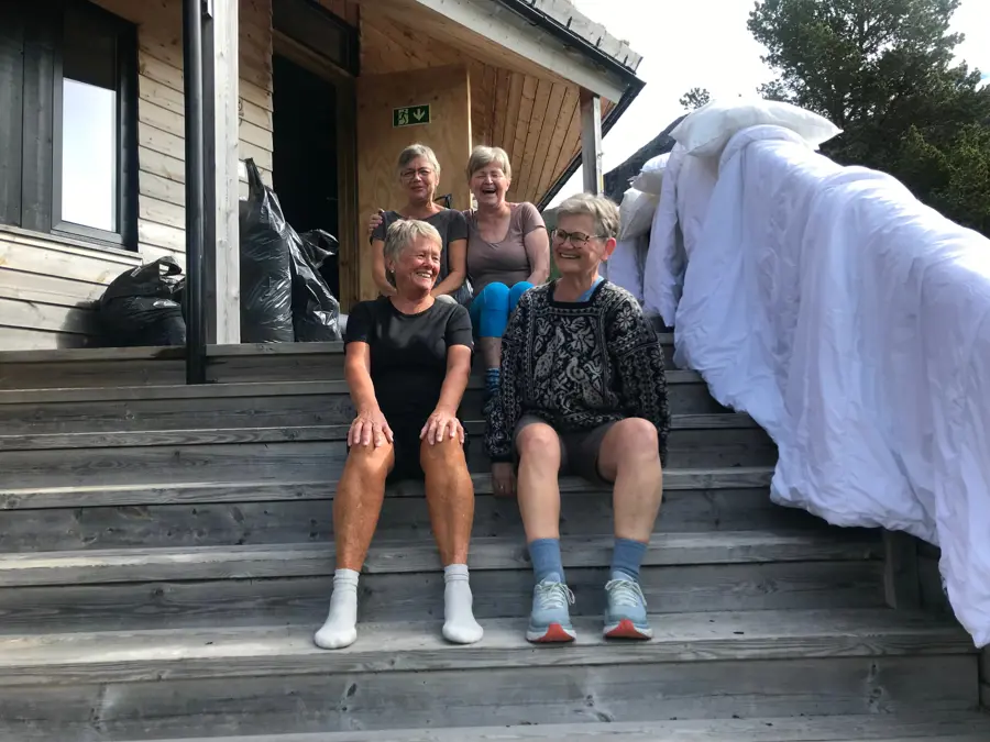Fire damer sitter på en trapp utenfor en grå hytte. Det henger dyner til lufting.