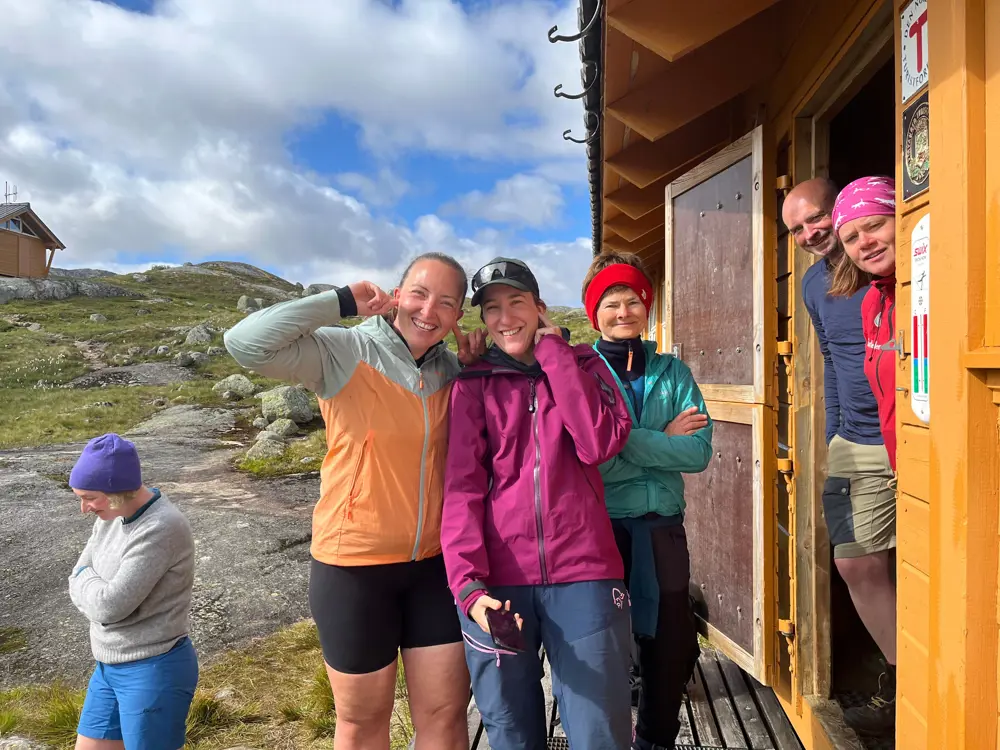 Et turfølge med menn og damer står utenfor en gul hytte og smiler til kamera.