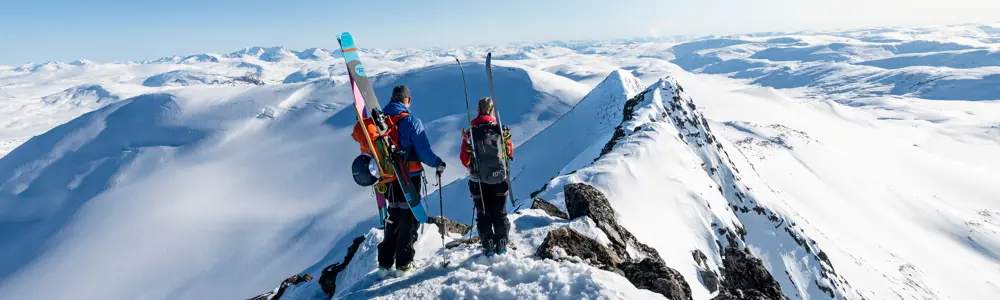 To personer står på en snødekt fjelltopp med skiutstyr, og ser utover et storslått vinterlandskap med fjell i horisonten.