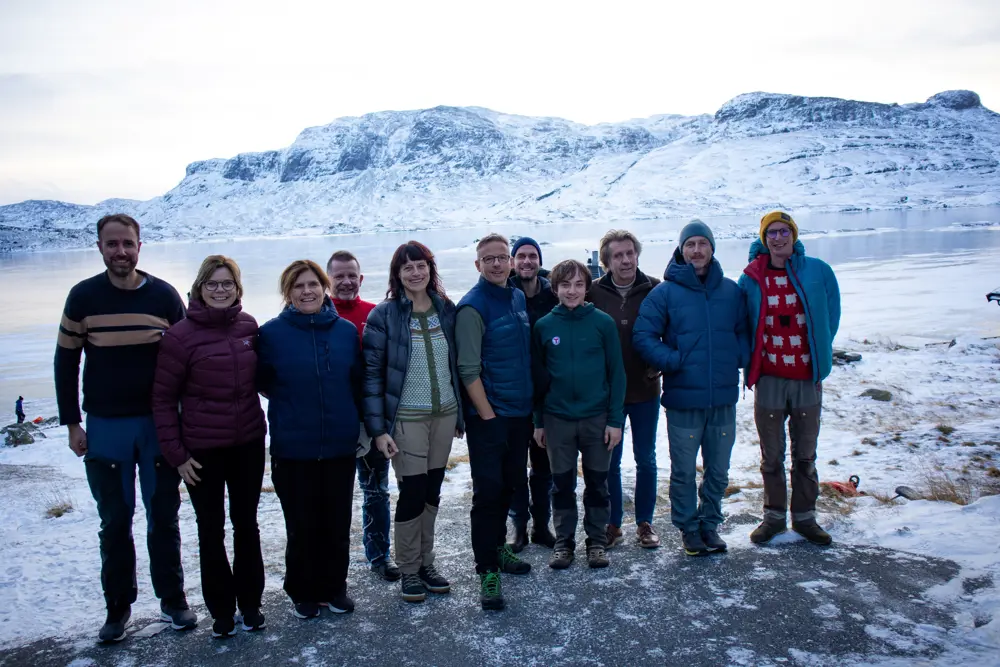 En gruppe med mennesker som står samlet og smiler til kamera. Bak dem er det fjell og vann som er is og snødekket.