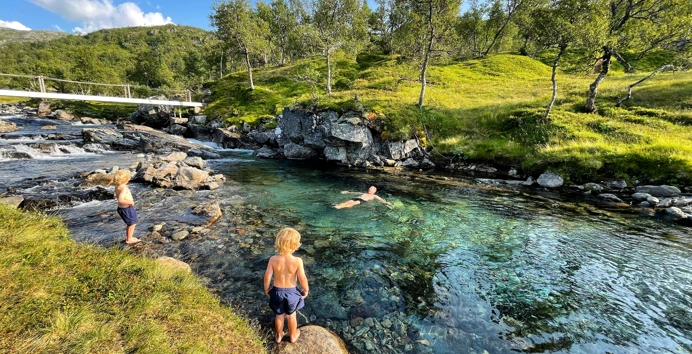 Bading i badekulp, en voksen mann bader og to unge gutter står ved elvekanten og ser på.