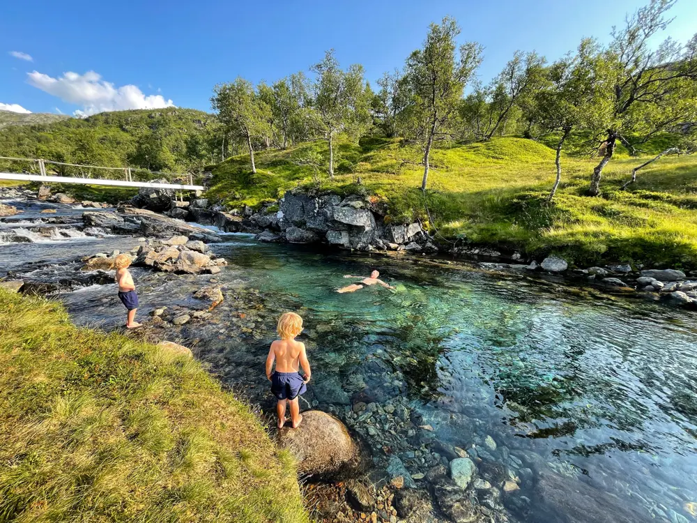 Bading i badekulp, en voksen mann bader og to unge gutter står ved elvekanten og ser på.