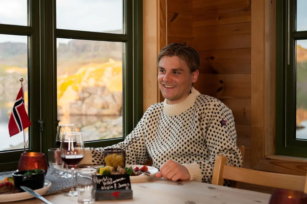 En mann sitter ved et bord og smiler. Foran han er det en tallerken med mat og et norsk flagg.