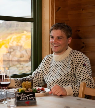 En mann sitter ved et bord og smiler. Foran han er det en tallerken med mat og et norsk flagg.