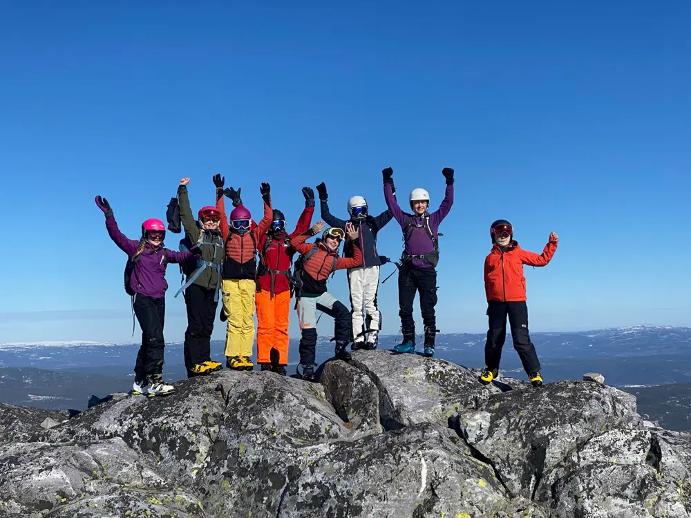 Ungdommer i skiutstyr står på toppen av et fjell med hendene i været
