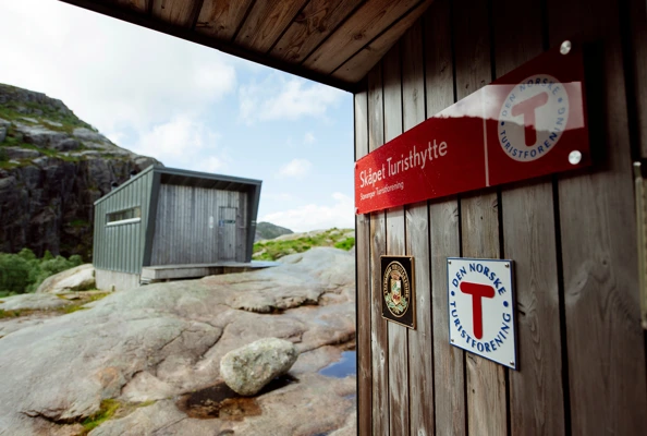 Bilde av et skilt med en T som henger på en grå hyttevegg.