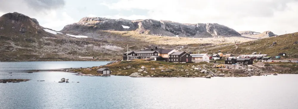 Dronebilde av en svartmalt hytte som ligger ved en innsjø og med fjell i bakgrunnen