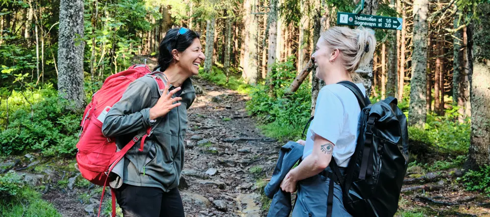 To personer smilende og pratsomme på en skogsti i Oslomarka. Begge bærer tursekker, og i bakgrunnen sees et skilt som viser retningene til Vettakollen (1,6 km) og Sognsvann (3,6 km)