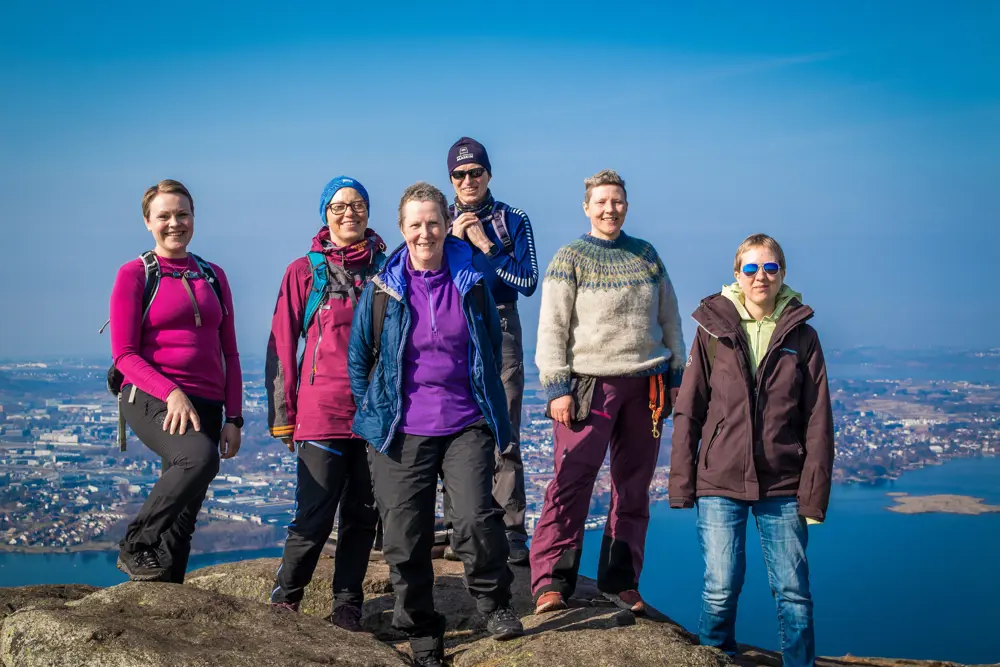 Seks personer står på toppen av et fjell og ser i kamera. Utsikten bak dem viser sjø og bebyggelse.