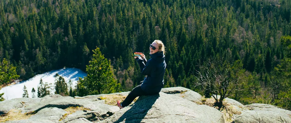 En kvinne sitter på en stein ute i naturen med en skogkledd åsside i bakgrunnen. Hun smiler mens hun holder en Kvikk Lunsj-sjokolade i hånden og har på seg solbriller og en mørk turjakke. Det ser ut som hun nyter en pause på en tur i skogen