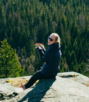 En kvinne sitter på en stein ute i naturen med en skogkledd åsside i bakgrunnen. Hun smiler mens hun holder en Kvikk Lunsj-sjokolade i hånden og har på seg solbriller og en mørk turjakke. Det ser ut som hun nyter en pause på en tur i skogen