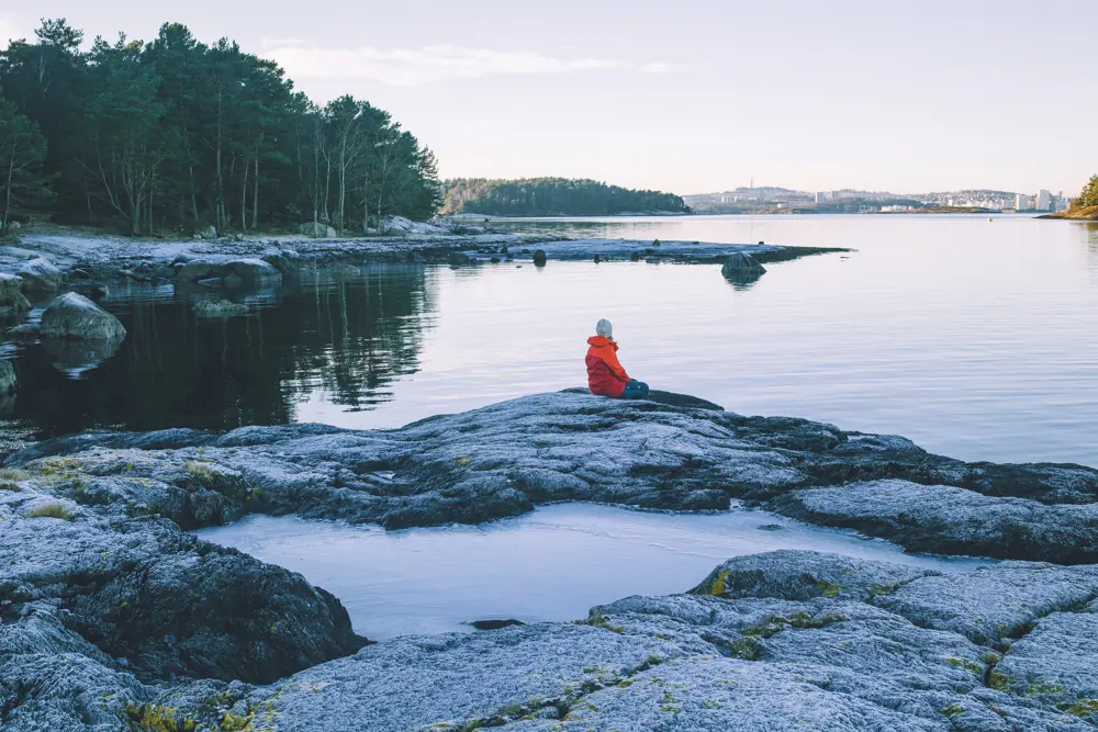 bilde av en person i oransj jakke som sitter på svaberg og ser utover vannet. 