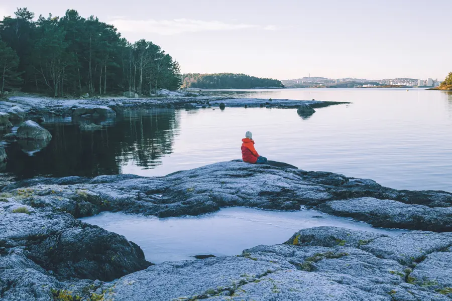 bilde av en person i oransj jakke som sitter på svaberg og ser utover vannet. 