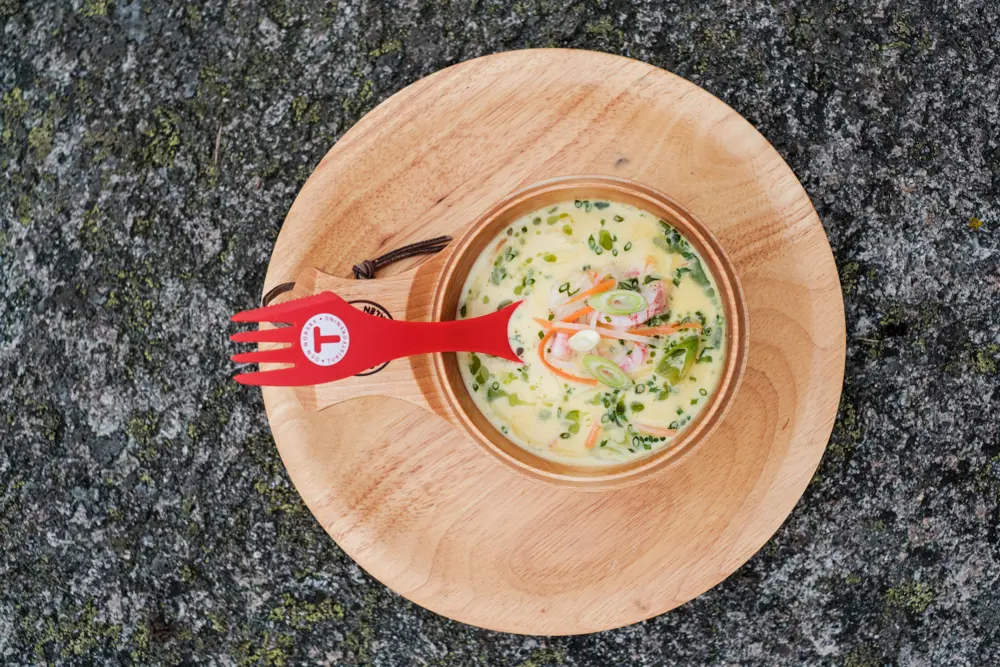 En tretallerken med en trekopp som inneholder fiskesuppe. Oppi suppa er det en rød skei med en T-logo på.