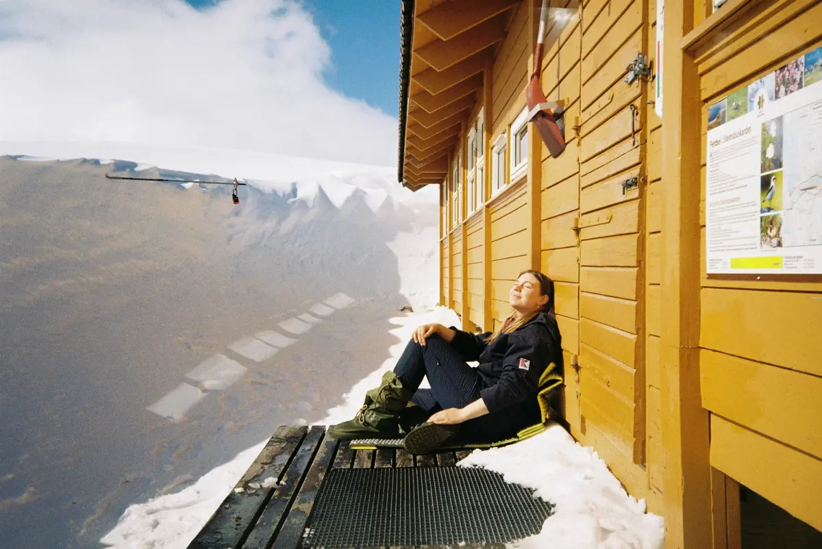 Ei jente sitter ned og nyter sola i hytteveggen på en gul hytte. Foran henne er det flere meter med brøytekant av snø.