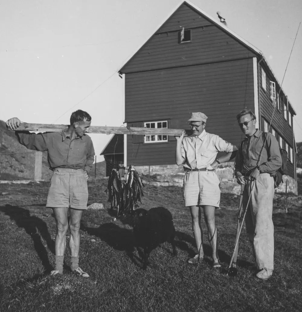 Gammelt bilde i svart-hvitt av tre menn som holder opp fisk foran hytte.