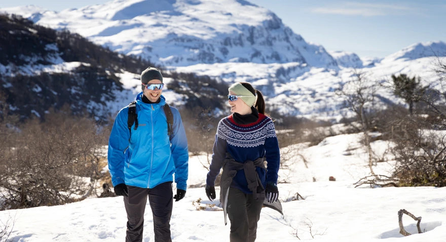 Mann i blå jakke og kvinne i mariusgenser smiler og går på tur i fjellet med snø og lyng.