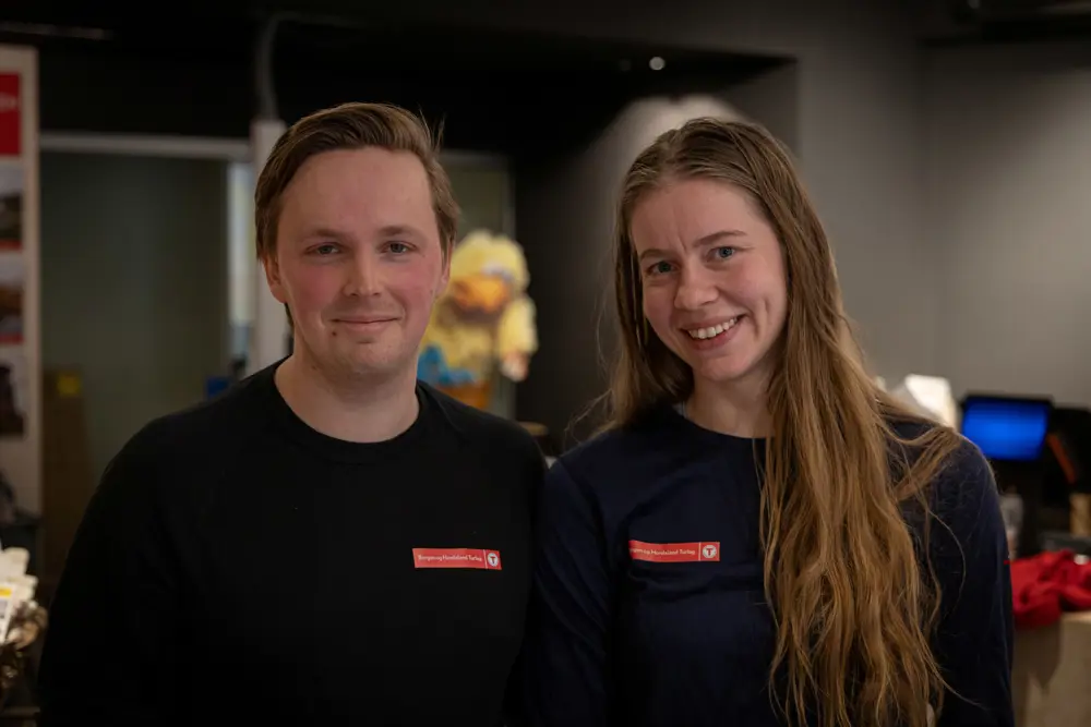 Mann og kvinne i T-merket genser smiler til kamera innendørs.
