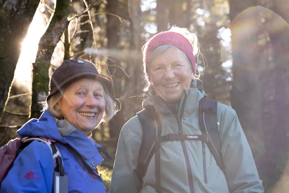 To voksne kvinner smiler til kamera når de er på tur i en skog.