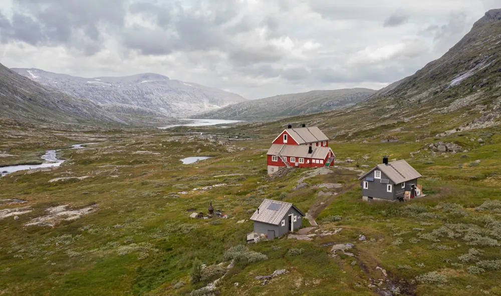 Rødmalt hytte i høyfjellet med to mindre blågrå hytter ved siden av.