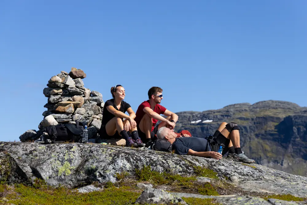 En kvinne og to menn sitter og slapper av ved en varde i solen på fjellet.