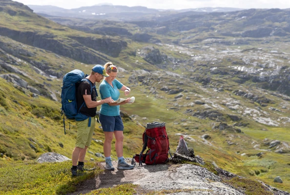 En mann og en dame i shorts og med ryggsekk studerer kart mens de er på tur i fjellet en sommerdag.