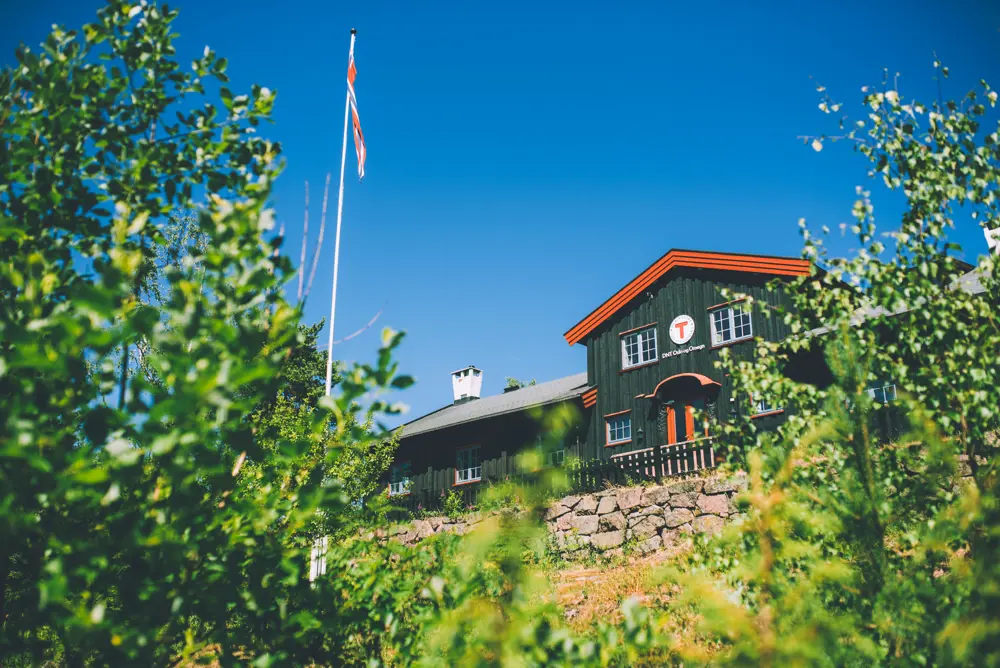 En svartmalt hytte med rødkarmer i solskinn. Det er grønne trær og busker i forkant. 