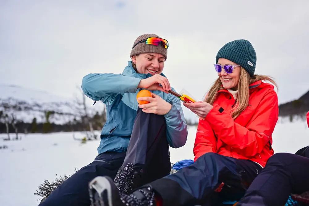 To kvinner sitter ute i vinterlandskap. De smiler og en holder en appelsin menst hun får sjokolade av den andre kvinnen. 