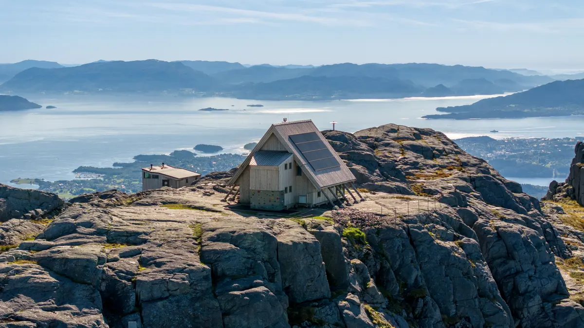 Hytte med skrått tak på toppen av fjellplatå med fjord bak.