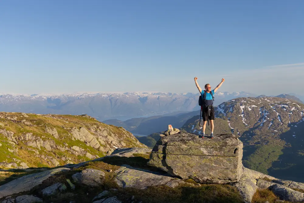 Mann 40+ står på stein med hendene i været, smiler, med Hardangerfjorden og fjell bak seg.