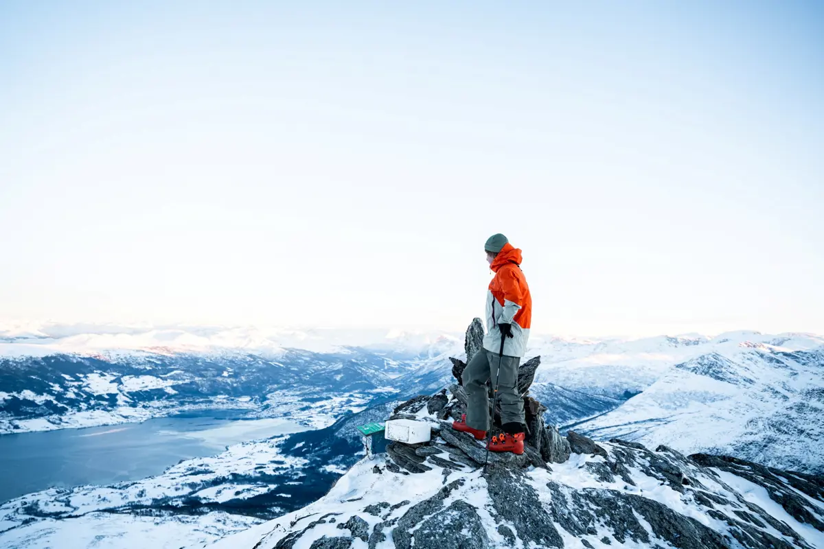 person i vinterklær og slalåmstøvler, står på en delvis snødekket fjelltipp og ser utover et fjell- og fjordlandskap.